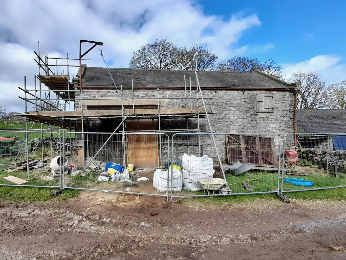 A stone barn under renovation with scaffolding, building materials and a skip in the yard