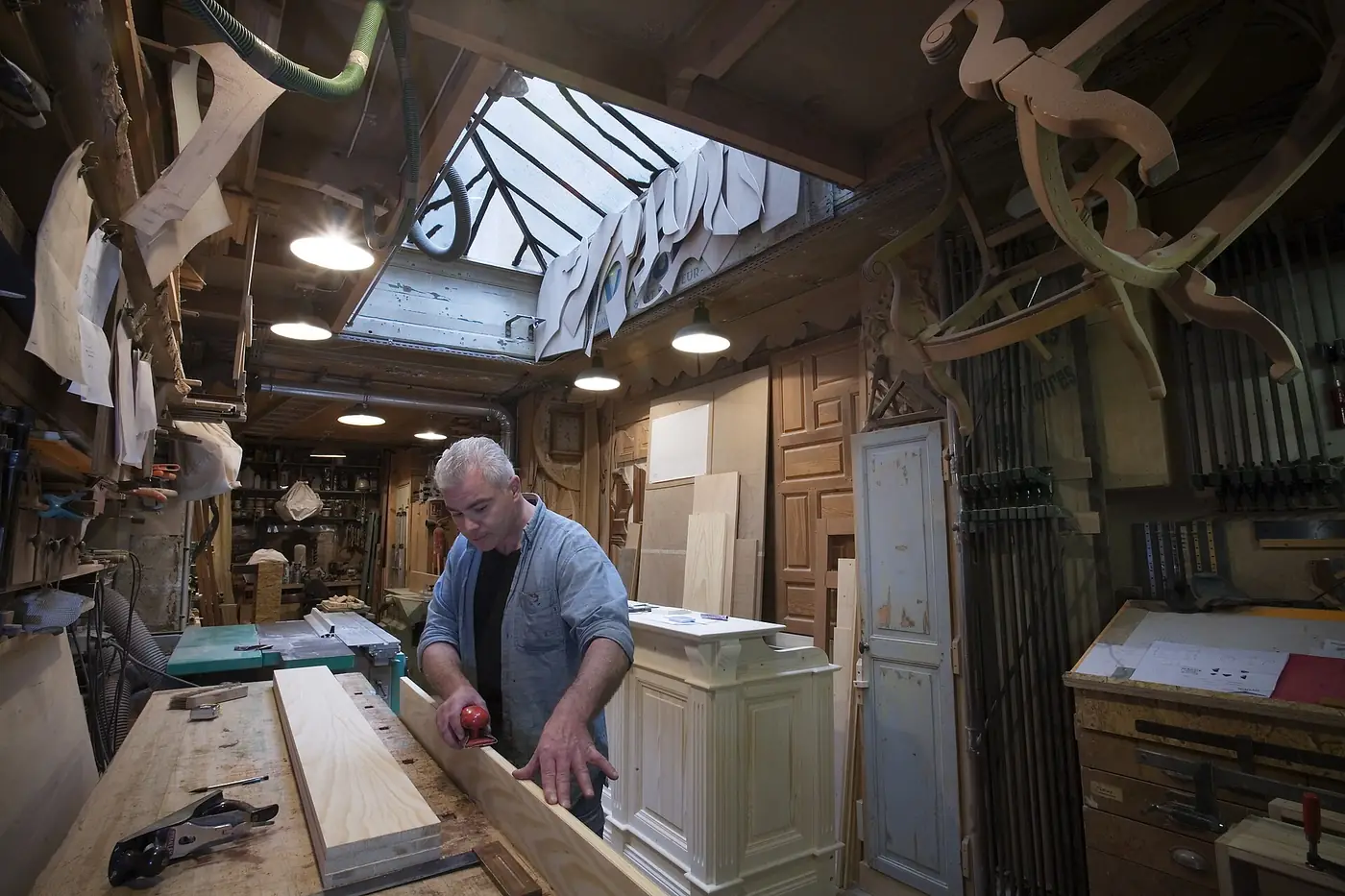 A carpenter planing timber at a workbench in a busy joinery workshop
