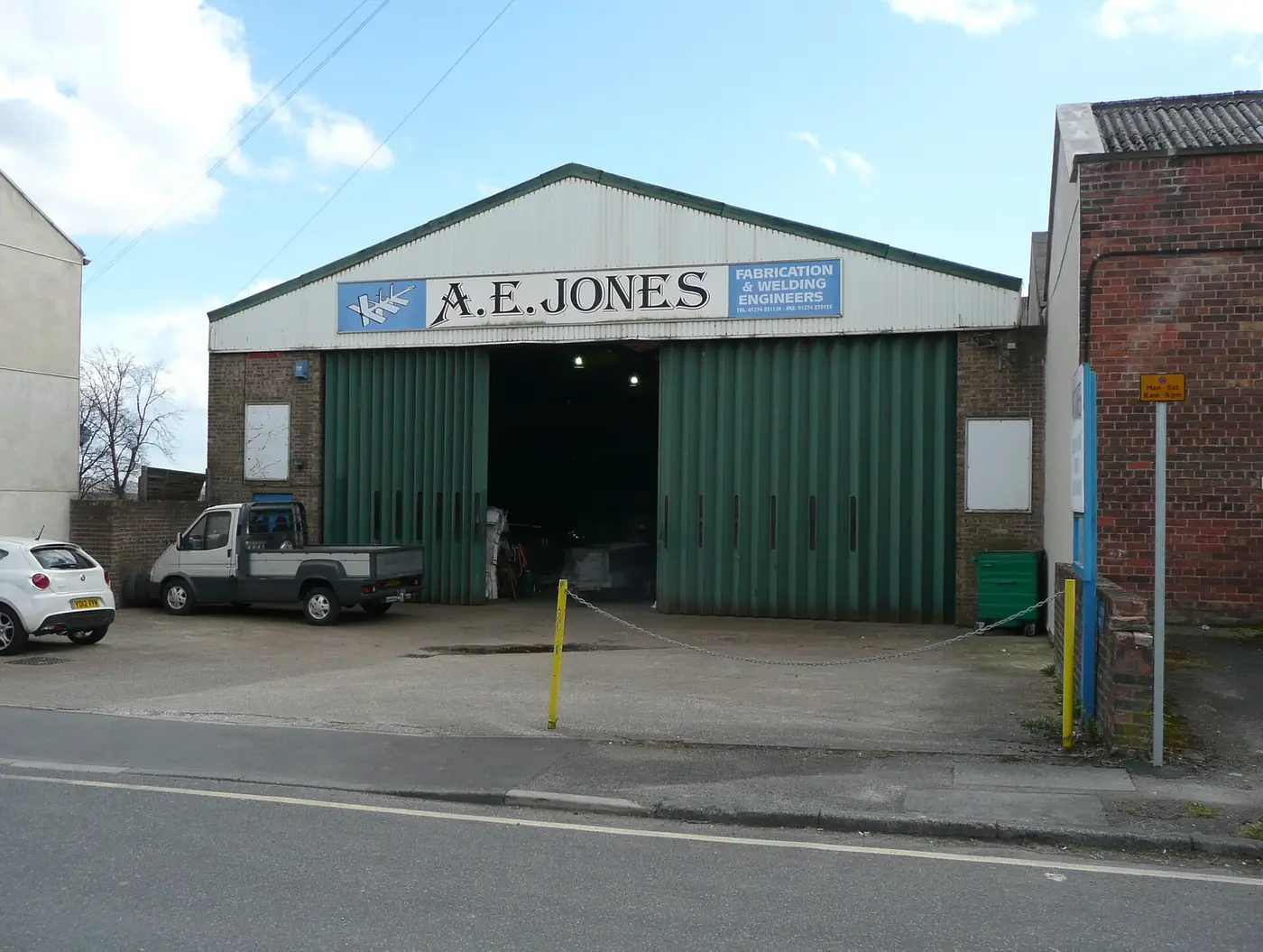 A metal fabrication workshop with a large green door, a sign above and vans parked outside