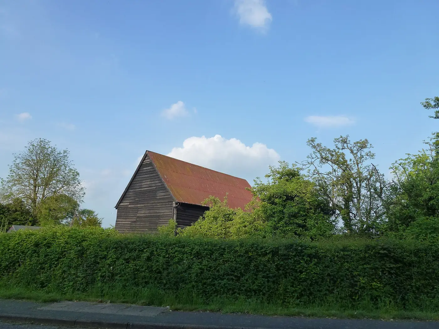 A black weatherboarded timber barn with a red roof behind a hedge